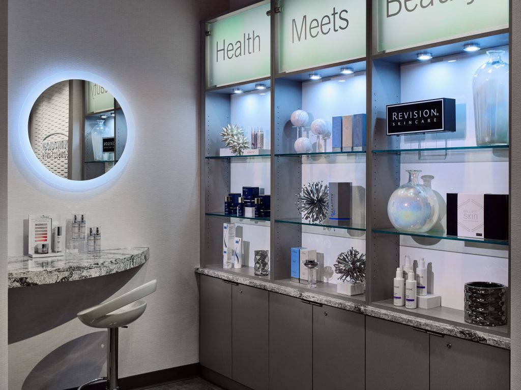corner of a dermatology office with a soft blue backlit round mirror and display shelves featuring different skincare products and decorative vases