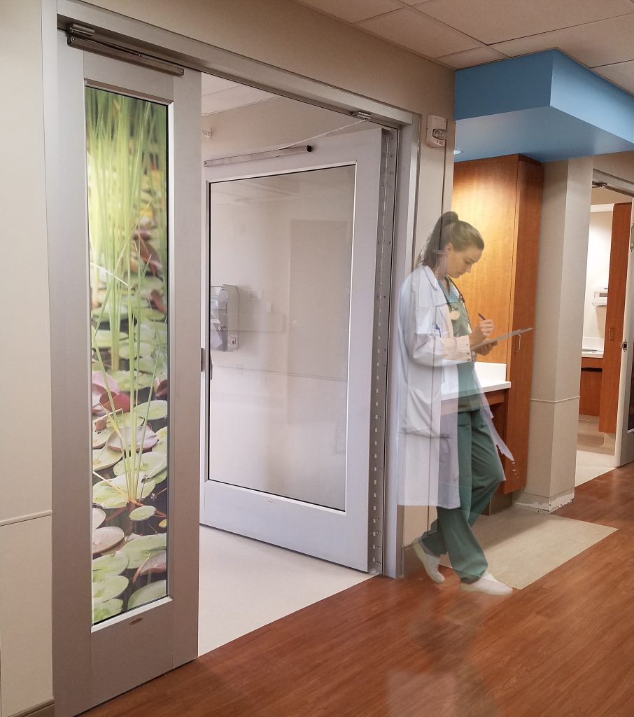 a woman in a lab coat leaning against a hospital hallway door writing on a clipboard