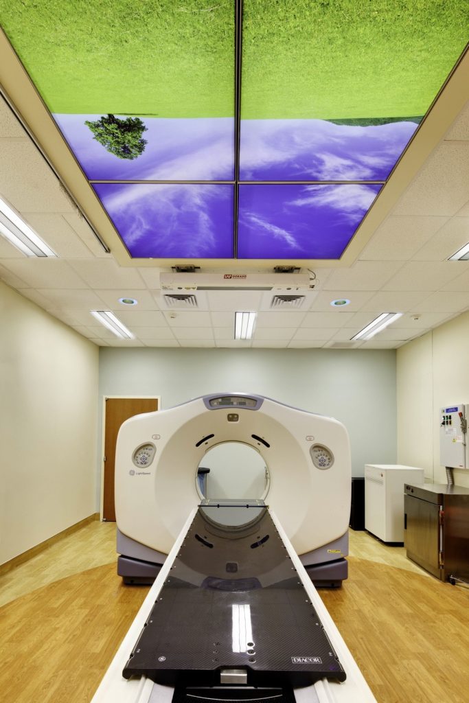 a CT scan machine in a room with decorative panels on the ceiling showing green grass, trees, and a deep blue sky