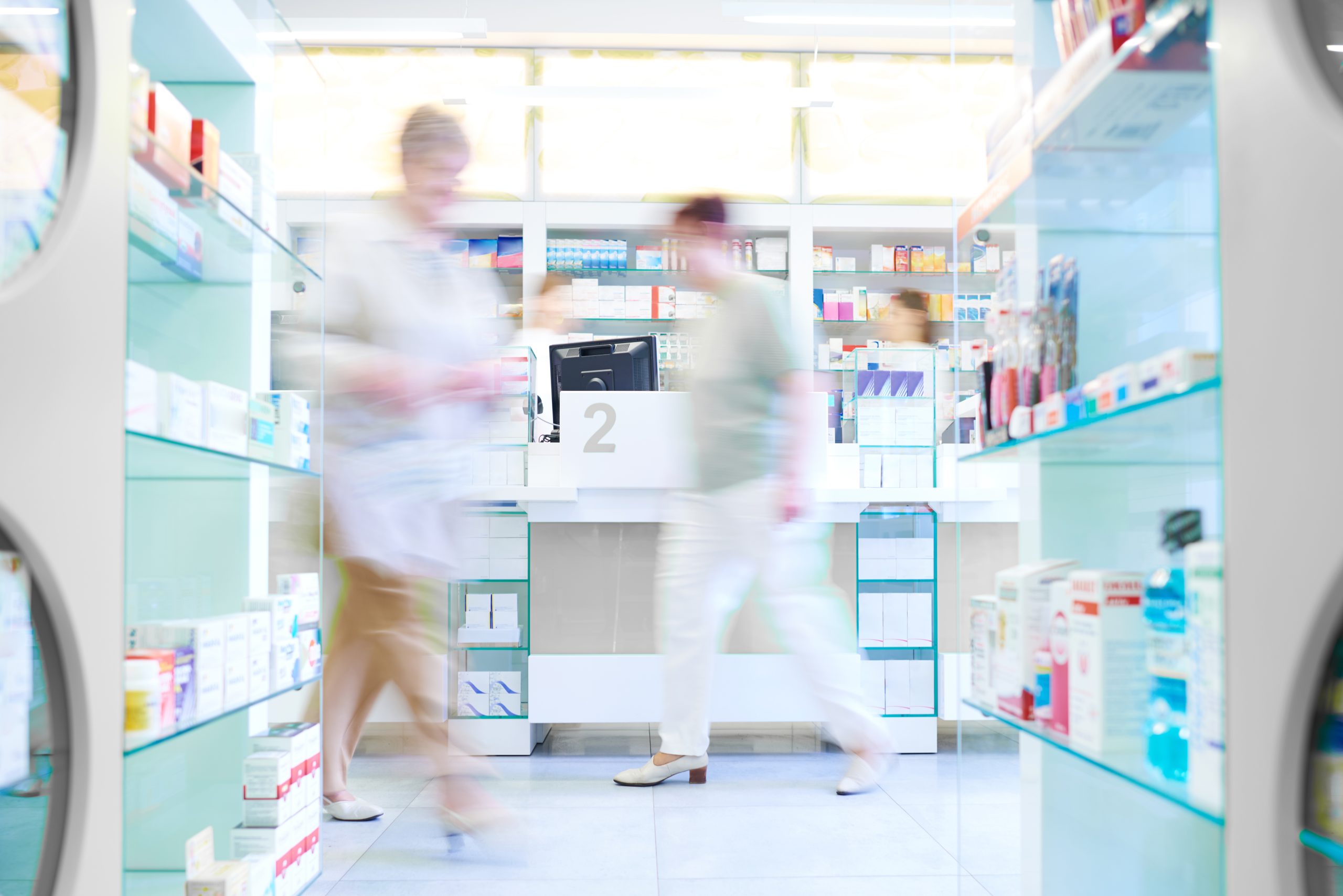 pharmacy with stocked shelves and two people blurred from walking through the aisles